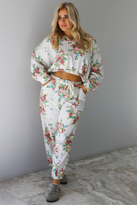 Woman wearing an oatmeal colored and multi-colored floral print two piece set featuring a collared long sleeved crop top and high waisted pants. The photo is taken indoors against a white wall with white marble flooring. 