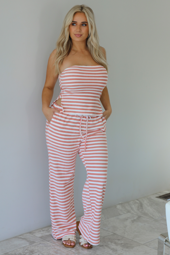 Woman wearing an orange, white, and green striped pattern set that has a strapless tube top and high waisted pants indoors against a white wall with white marble flooring. 