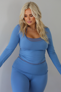 A woman modeling a matching two-piece lounge set in a dusty blue color, featuring a long-sleeve scoop-neck top with a connected darker blue halter neck detail and matching high-waisted flared pants, standing indoors against a white wall. The photo shows a closeup of the top and waistline of the pants. 