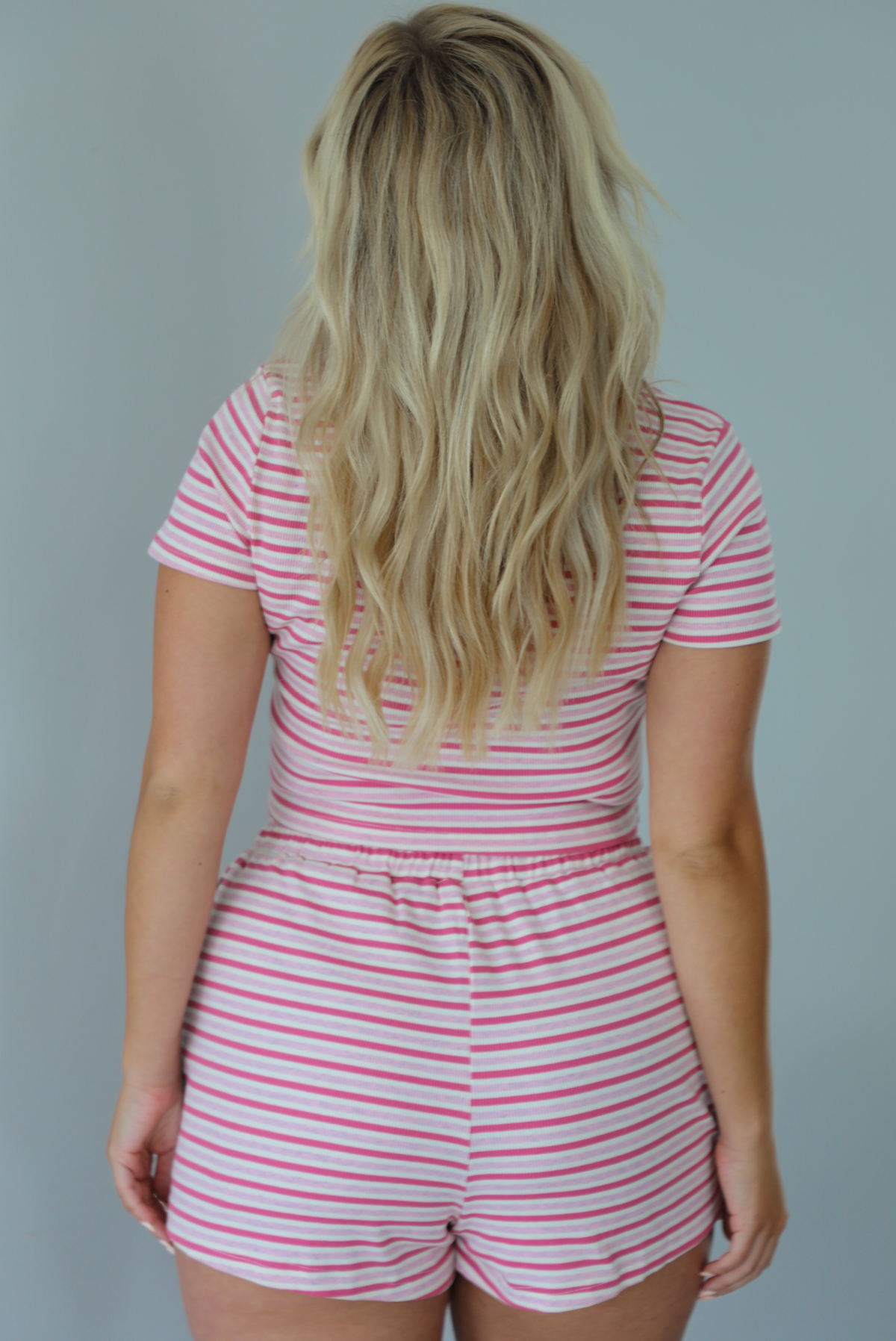 Woman wearing a pink and multi-colored striped patterned two piece set featuring a cropped t-shirt and high waisted, elastic waist shorts indoors against a white wall and shows the back of the set. 