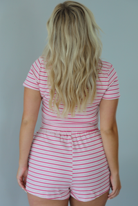 Woman wearing a pink and multi-colored striped patterned two piece set featuring a cropped t-shirt and high waisted, elastic waist shorts indoors against a white wall and shows the back of the set. 