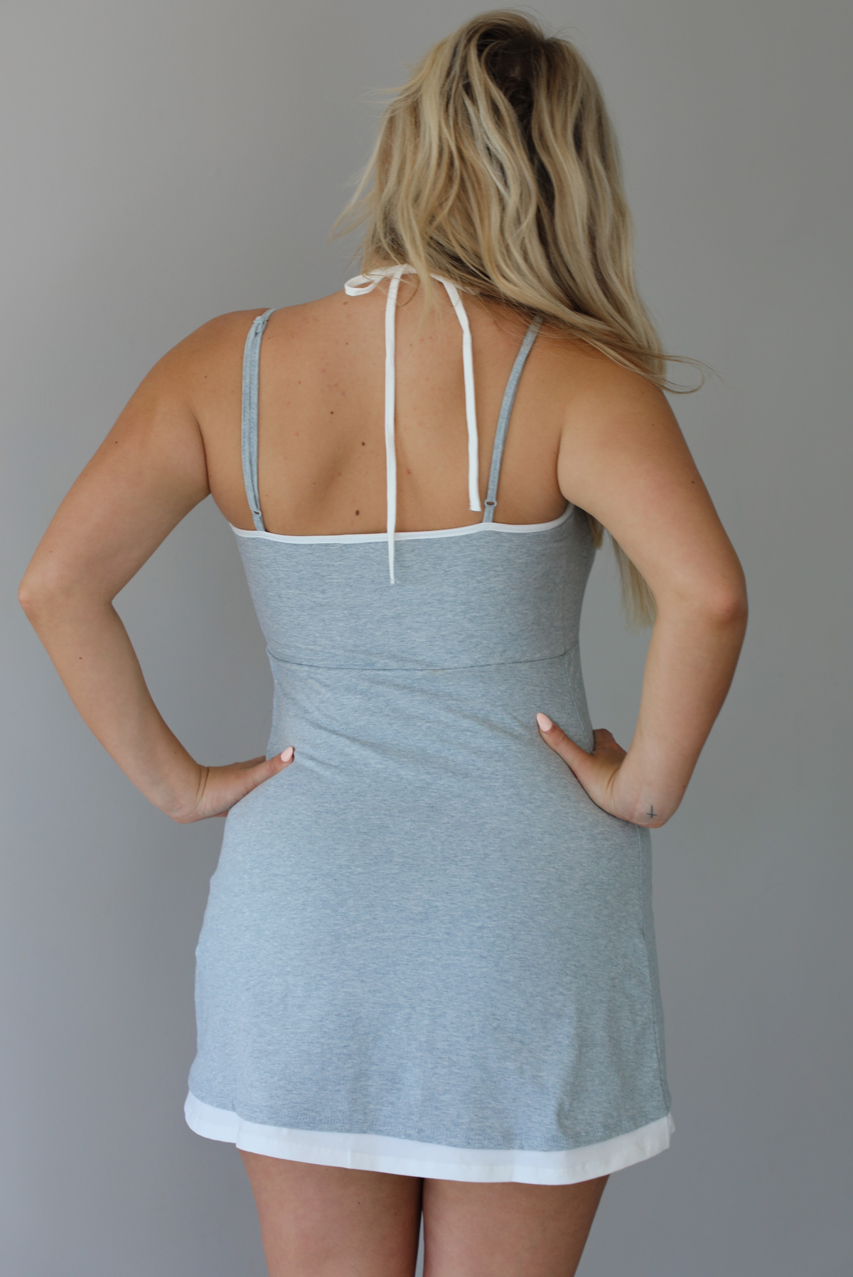 Woman wearing a heather grey athleisure dress with white trim detailing at the hem indoors against a white wall. The photo shows the back of the dress, along with the adjustable straps, and the halter tie neckline. 