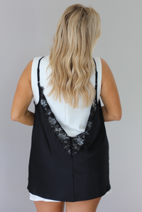 Woman wearing a white mini-length dress that has a black lace tank detail overlay. The photo is taken indoors against a white wall and shows the back of the dress. 