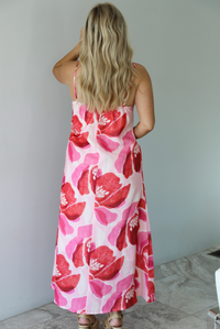 Woman wearing a pink and red floral print maxi dress with thin straps and a flowy fit indoors against a white wall with white marble flooring. The photo shows the back of the dress. 
