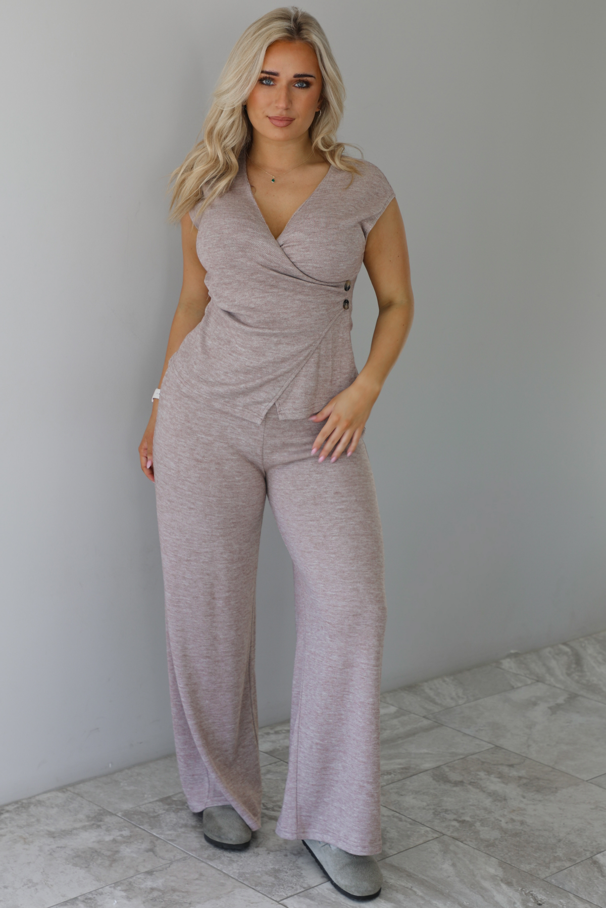 Woman wearing a taupe colored two piece set featuring a vest-appearance top with non-functional button closures on the left side and high waisted pants. The photo is taken indoors against a white wall with white marble flooring. 