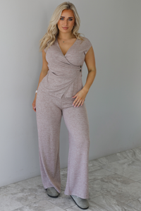 Woman wearing a taupe colored two piece set featuring a vest-appearance top with non-functional button closures on the left side and high waisted pants. The photo is taken indoors against a white wall with white marble flooring. 
