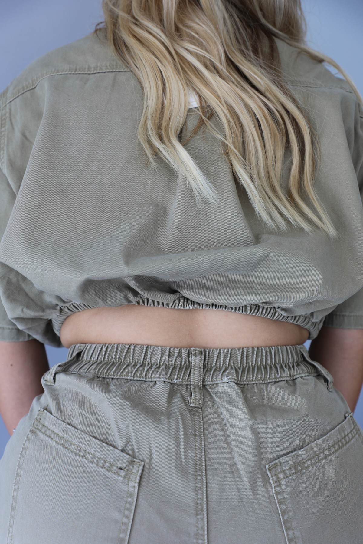 A closeup of a woman modeling a short-sleeve khaki jumpsuit with a button-front collared romper with a cinched waist, standing indoors against a white wall. It shows the cut out and elastic waistline in the back. 