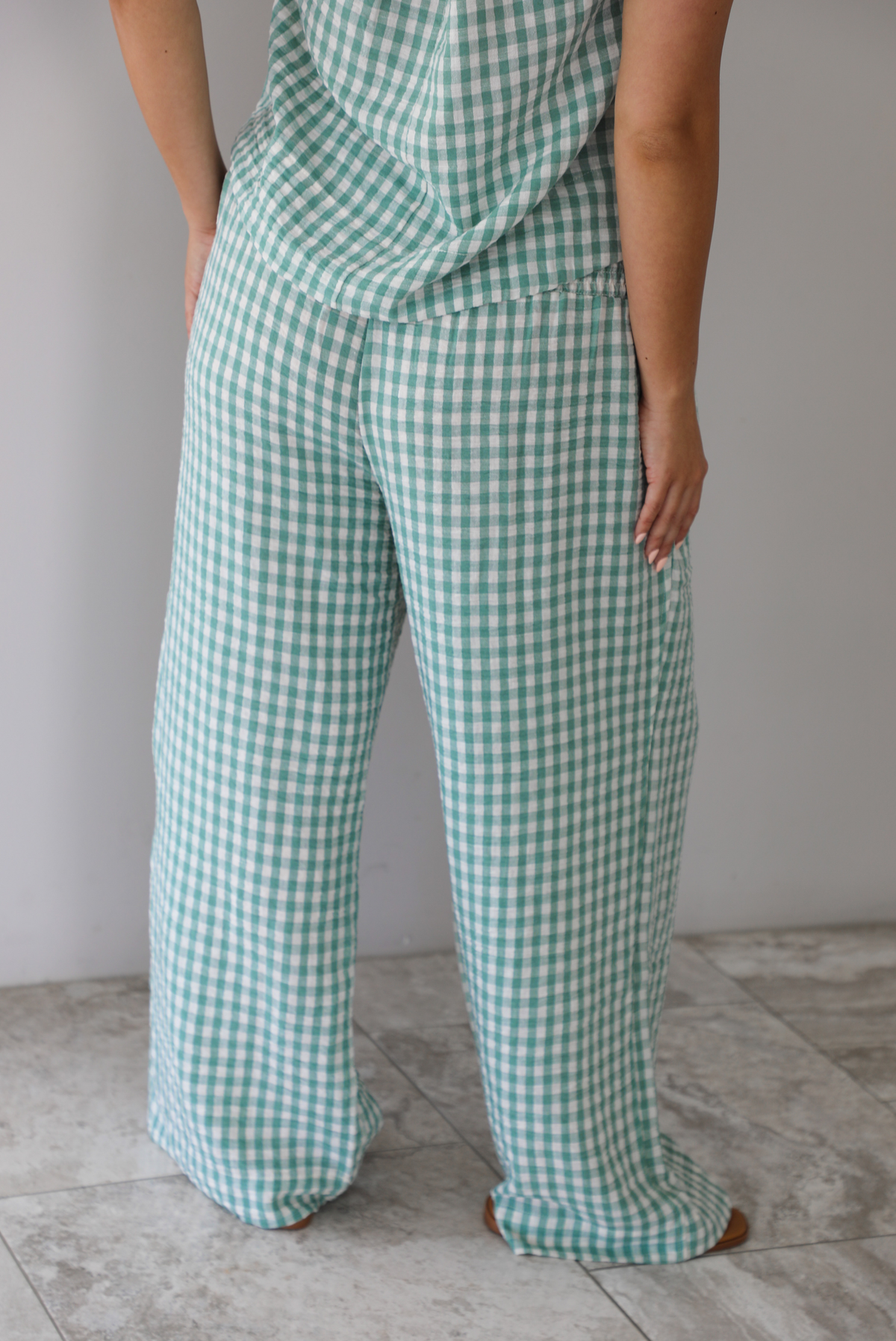 Woman wearing a mint and white gingham set with an oversized top and flared hem pants indoors against a white wall with white marble flooring. The photo shows a closeup of the back of the pants, which have a smocked waistline and functional pockets on either side. 