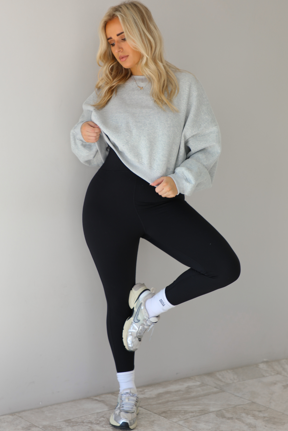 Woman wearing a heather grey cropped sweatshirt with a black fitted jumpsuit underneath. The outfit is paired with high ankle socks and silver sneakers. The photo is taken indoors against a white wall with white marble flooring. 