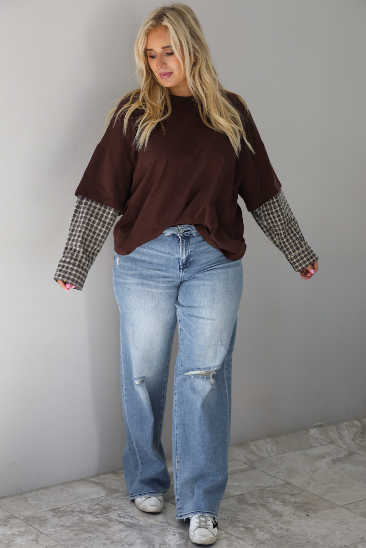 A woman wearing a brown t-shirt with a layered look of plaid sleeve detail, paired with light wash denim jeans, standing indoors against a white wall with white marble flooring. 