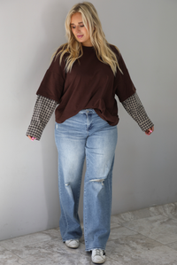 A woman wearing a brown t-shirt with a layered look of plaid sleeve detail, paired with light wash denim jeans, standing indoors against a white wall with white marble flooring. 