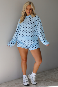 A woman wearing a blue two piece set with a navy polka dot pattern throughout, featuring a sweatshirt and high waisted sweat-shorts, paired with chunky sneakers and high ankle socks, shown indoors against a white wall with white marble flooring. 