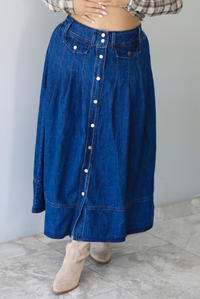 A woman wearing a long blue denim button-front skirt with a plaid long-sleeve top and beige ankle boots, standing indoors against a white wall with white marble flooring. 