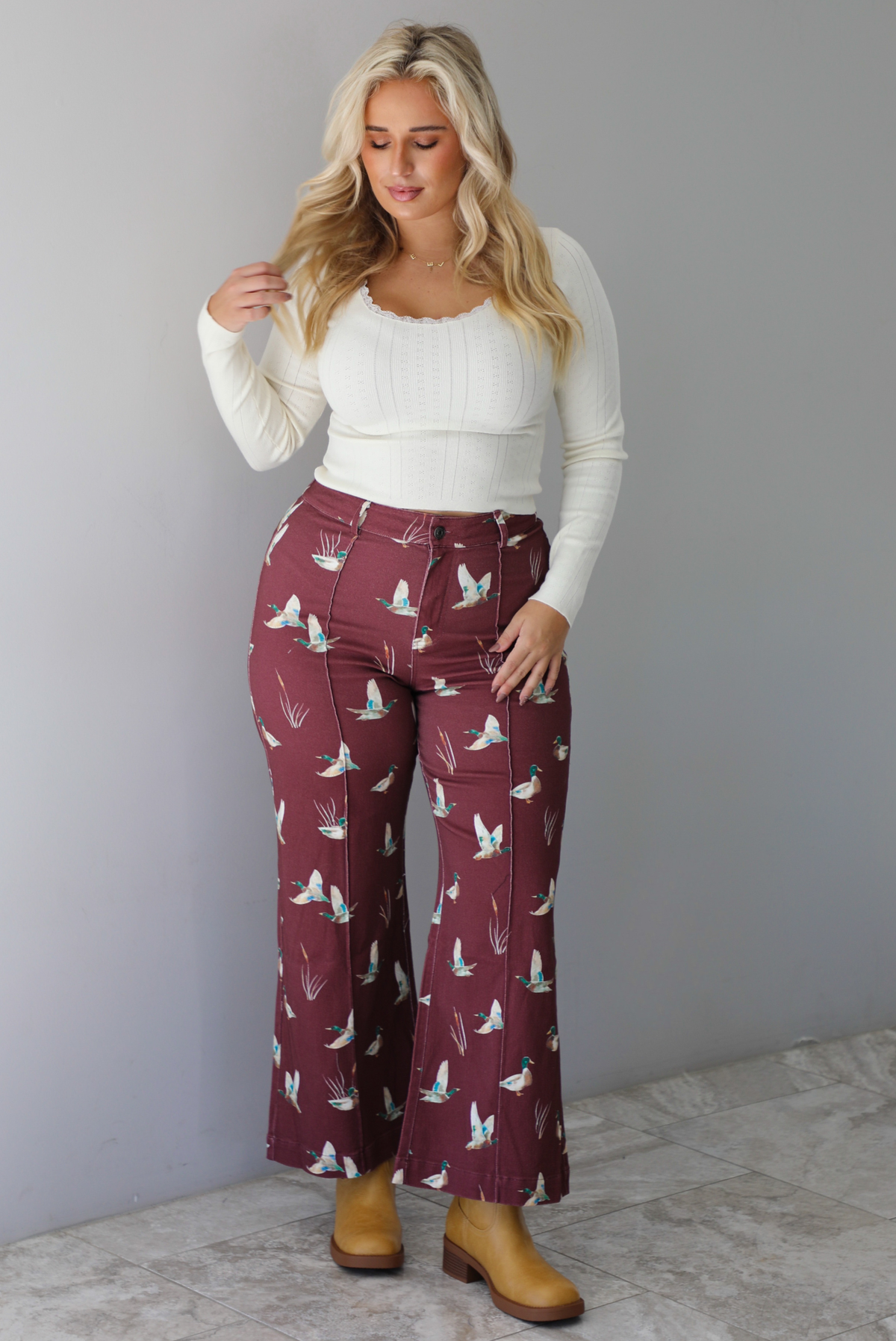 A woman wearing a cream top and maroon high waisted pants with a repeating mallard print throughout, standing indoors against a white wall with white marble flooring. 