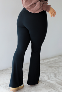 Woman wearing black leggings that have a high waistline with a folded over detail with a slightly flared hem. The photo shows the back of the leggings. The photo is taken in front of a white wall with white marble flooring. 