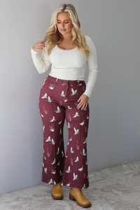 A woman wearing a cream colored knit long sleeved top paired with high waisted maroon jeans with a repeating mallard design, standing indoors against a white wall with white marble flooring. 