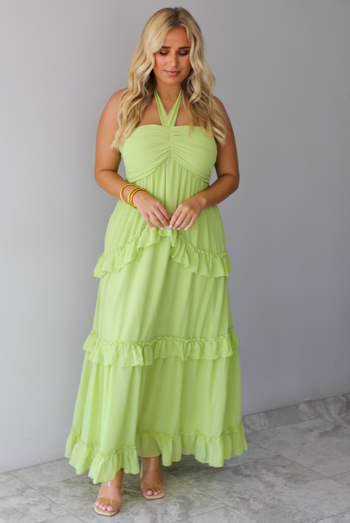 A woman modeling a lime green halter-neck maxi dress with tiered ruffle details, standing indoors against a white wall with white marble flooring. 