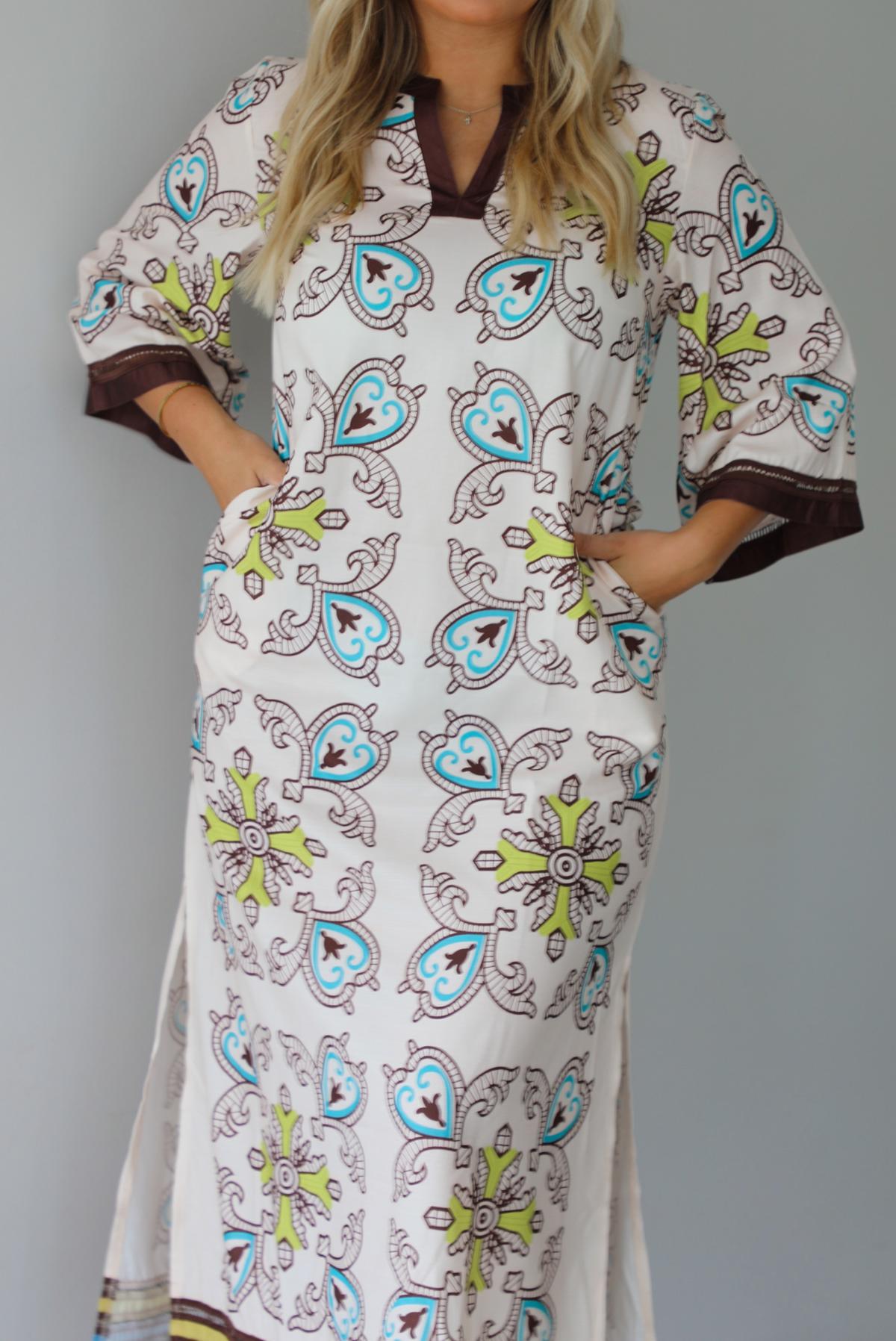 A woman modeling a long kaftan dress with a colorful paisley pattern on a cream background, featuring brown trim and sandals, standing indoors against a white wall.