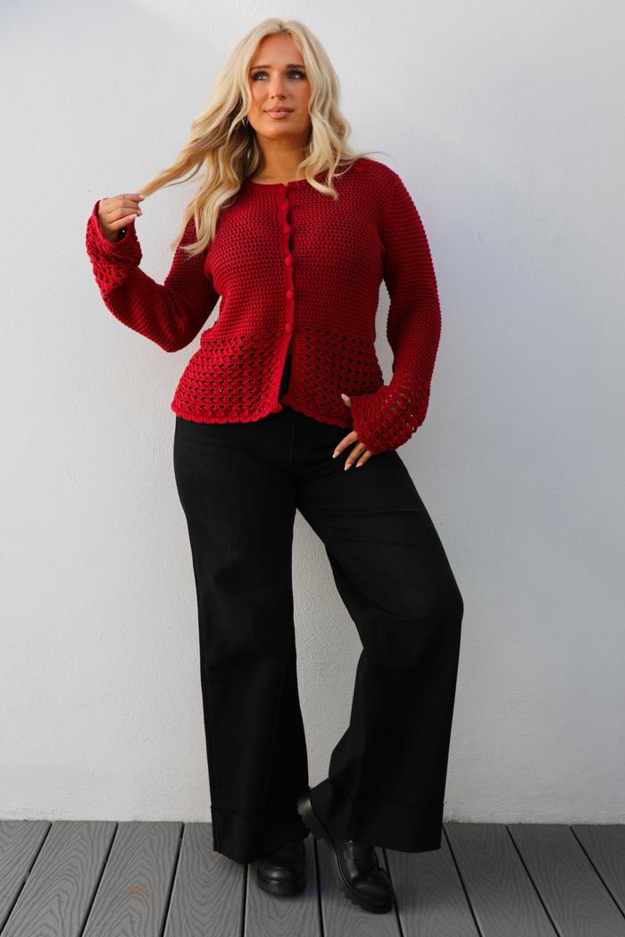 Woman model wearing a burgundy knit cardigan with button closures in the front and black denim jeans. The photo is taken outdoors against a white wall. 