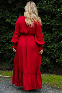 Woman wearing a red maxi length dress that has a v-neckline with button closures at the bodice, a cinched with elastic waistline, long sleeves, and a flowy skirt. The photo is taken outdoors against greenery. Photo shows the back of the dress. 