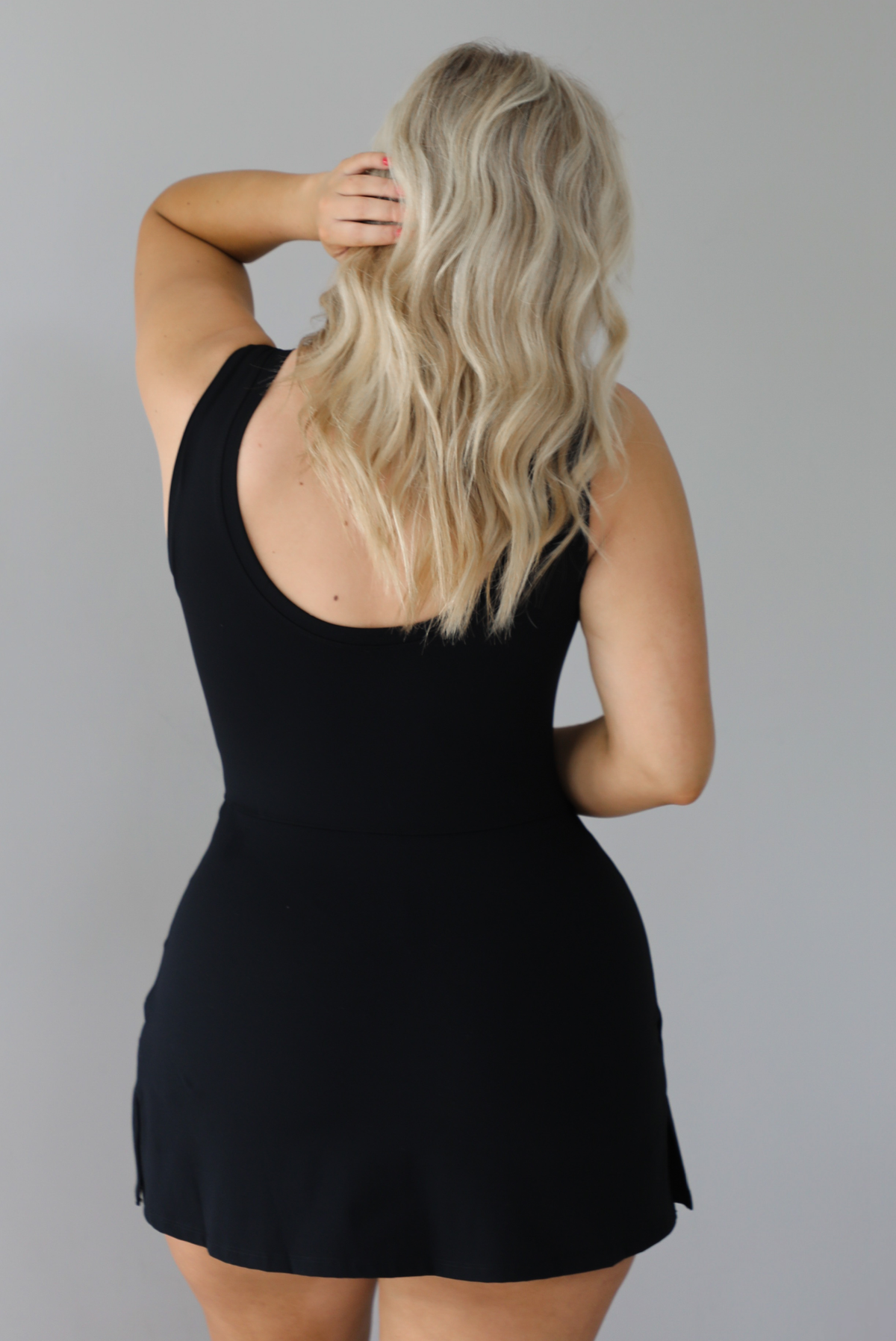Woman wearing a black athleisure romper that has a wrap-style skirt detail that wraps around and ties in the front on the left side. The photo shows the back of the romper that has a scooped design in the back and looks like a dress. The photo is taken indoors against a white wall with white marbled flooring. 