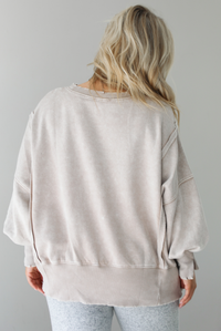 Woman wearing sand colored oversized sweatshirt with exposed seam details and a high-low design with heather grey casual pants. The photo shows the back of the sweater. The photo is taken indoors against a white wall.