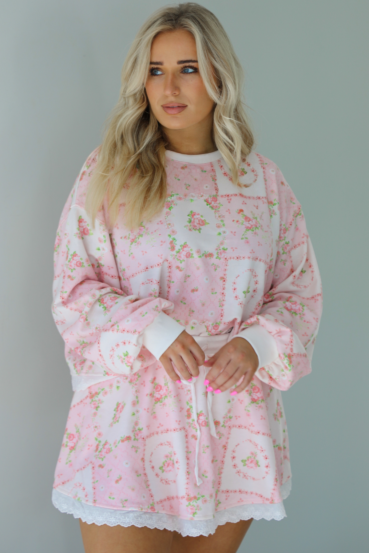 Woman wearing a two piece set featuring a pink floral set that has an oversized long sleeved top and high waisted skirt. The photo is taken indoors against a white wall.
