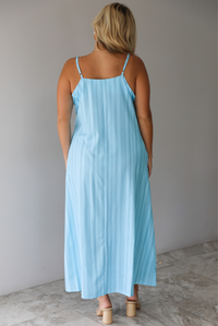 A woman shown from behind modeling a light blue sleeveless maxi dress with vertical grey pinstripes and side pockets, standing indoors against a white wall with white marble flooring. 