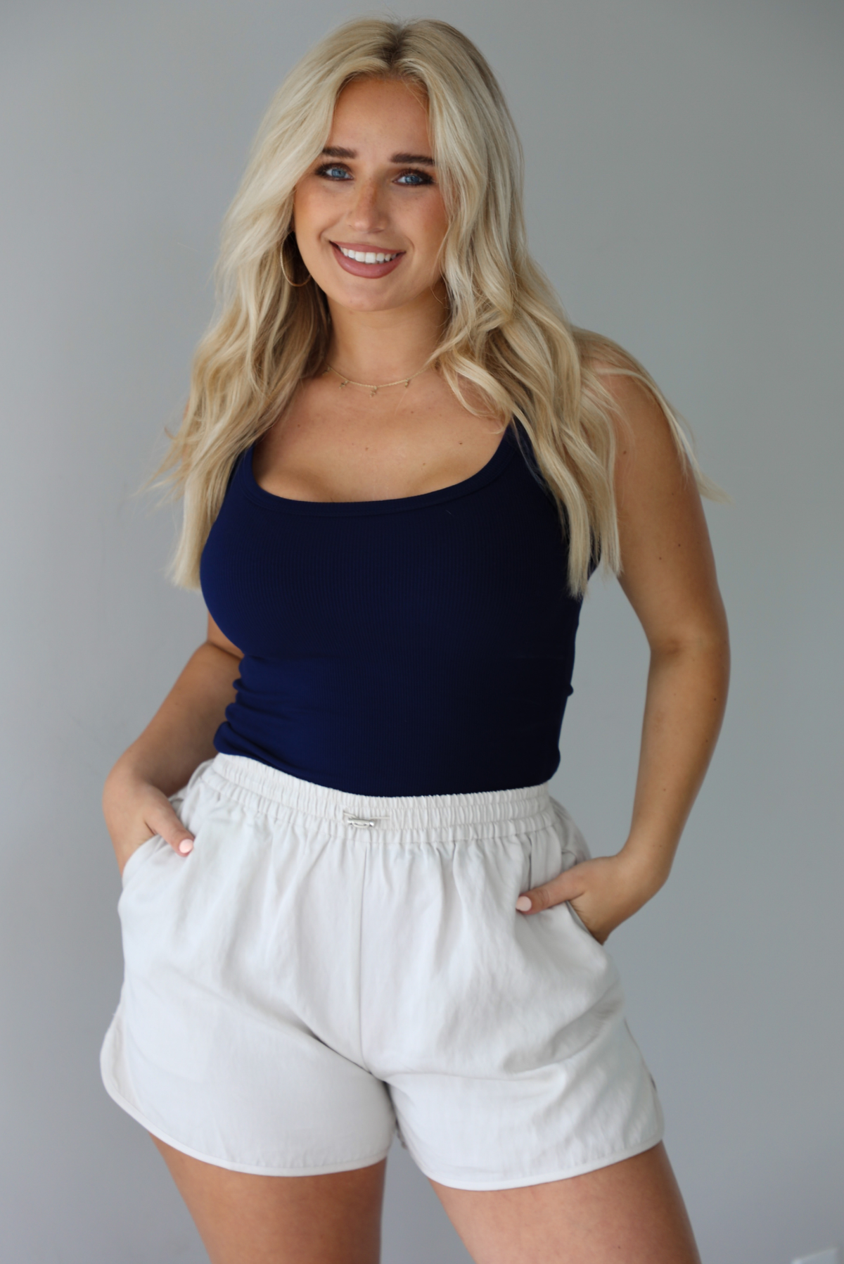Woman wearing a navy ribbed tank tucked into stone colored shorts indoors against a white wall. 
