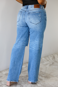 Woman wearing a pair of medium-wash, mid-rise denim jeans that have distressed details in the front and a flared, boot-cut-like hem. The photo is taken indoors against a white wall with white marble flooring and shows the back of the jeans. 