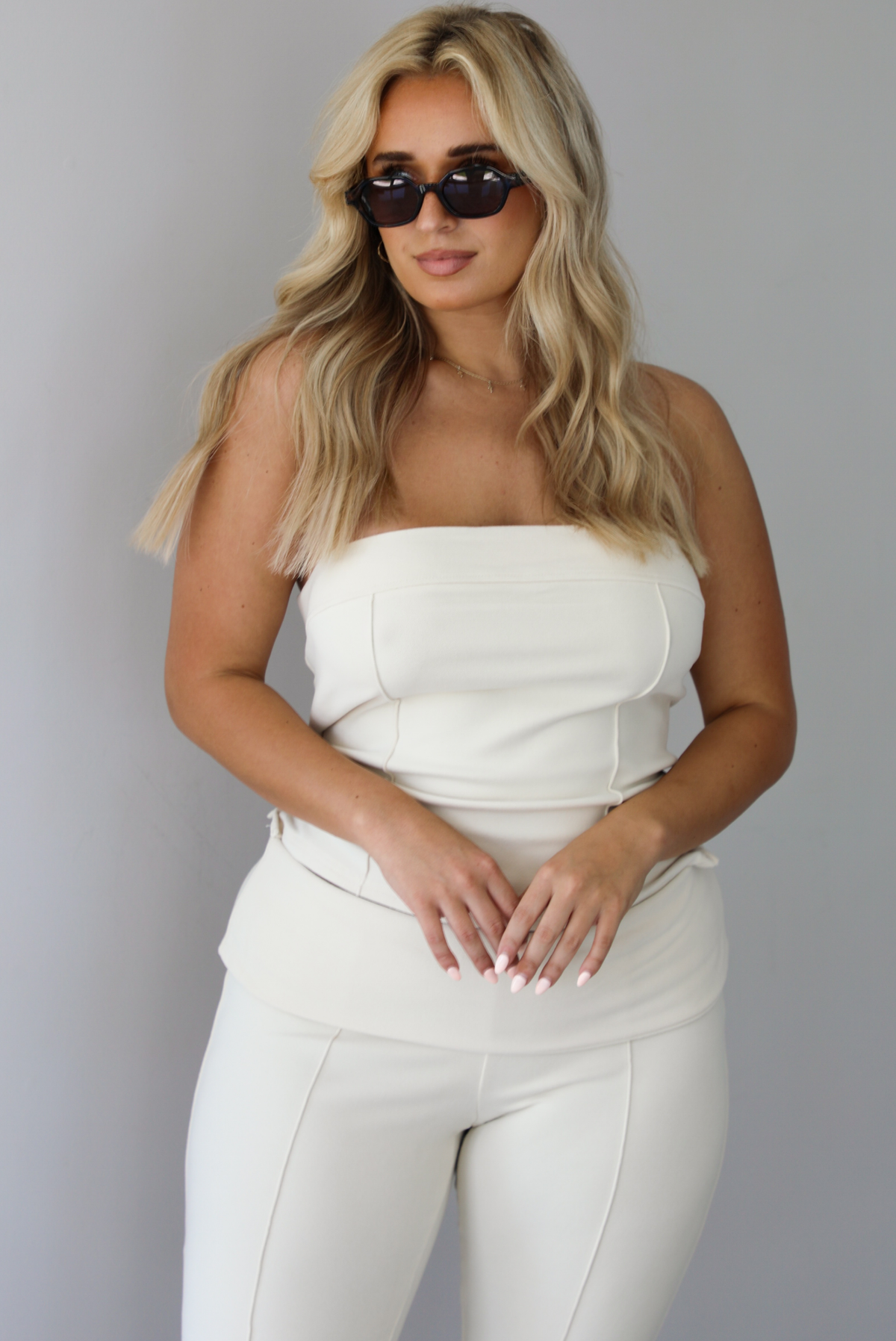 Woman wearing a cream two piece set featuring a strapless tube top and capri pants that fold over at the waist indoors against a white wall. The photo shows a closeup of the tube top, which has seam details. 