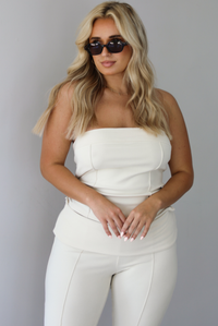 Woman wearing a cream two piece set featuring a strapless tube top and capri pants that fold over at the waist indoors against a white wall. The photo shows a closeup of the tube top, which has seam details. 