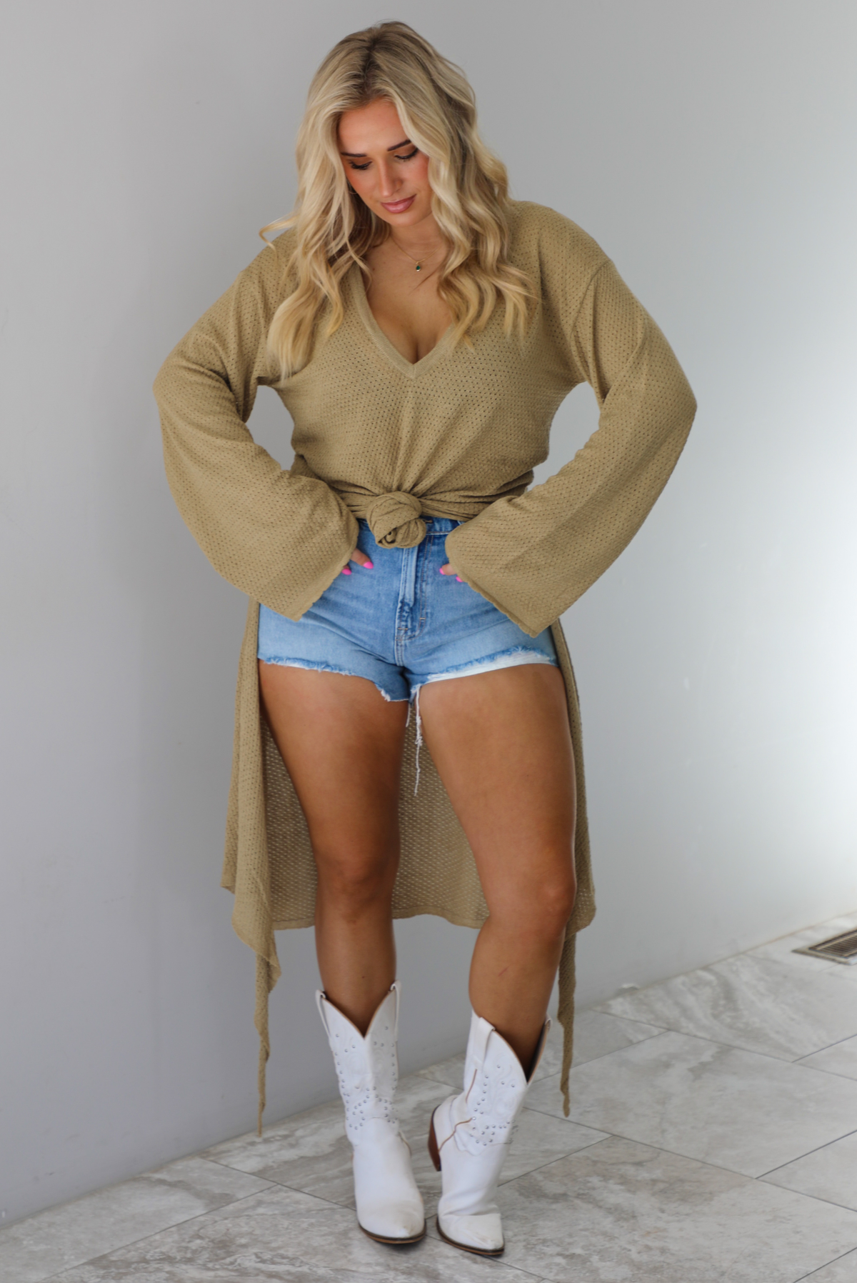 Woman wearing a camel long-sleeve top that's tied in the front with a knot and has a train in the back. The top is paired with denim shorts and white cowboy boots. The photo is taken indoors against a white wall with white marble flooring. 