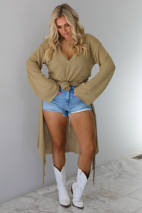 Woman wearing a camel long-sleeve top that's tied in the front with a knot and has a train in the back. The top is paired with denim shorts and white cowboy boots. The photo is taken indoors against a white wall with white marble flooring. 