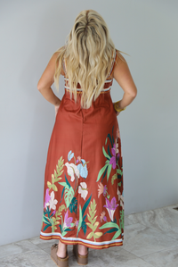 A woman shown from behind wearing a rust colored maxi dress with other colors at the bust in a striped pattern, with a multi-colored floral pattern at the hem, standing indoors against a white wall with white marble flooring. 