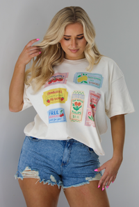 Woman wearing a white relaxed fit graphic tee that has various tickets on it with different fruits and flowers on them. The tee is tucked into a pair of distressed denim shorts. The photo is taken indoors against a white wall.