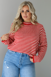 Woman wearing a red and white striped sweater with high waisted medium wash flared hem denim. The photo shows a closeup of the sweater which has a front tuck. The photo is taken in front of a white wall.