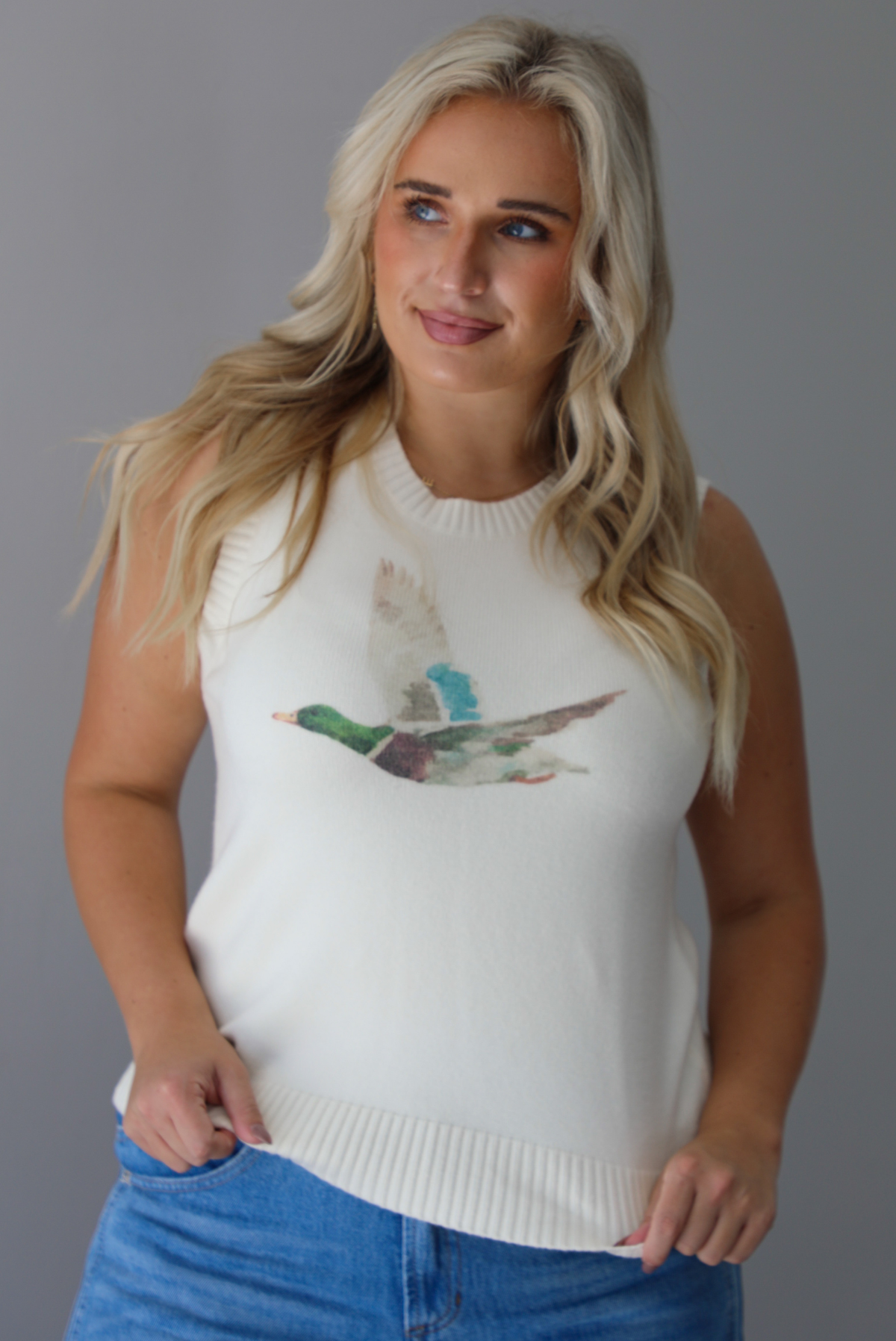 A woman wearing a cream colored sweater vest with a mallard duck on the front, paired with medium wash denim with wide leg hem, standing indoors against a white wall.