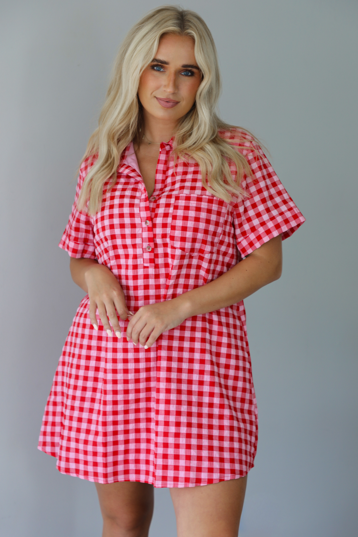 Woman wearing a pink and red gingham collared neckline t-shirt dress indoors against a white wall. The photo shows a closeup of the dress, and showcases the button closures at the neckline. 
