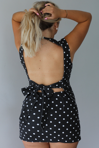 Woman wearing a black set with white polka dots denim set with black boots indoors against a white wall. The photo shows the back and how the top has an open back and tie closure. 