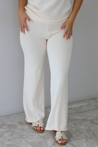 Woman wearing a cream two piece set featuring a high-neck sleeveless top and matching mid-rise pants indoors against a white wall with white marble flooring. The photo shows a closeup of the pants, which have a raw, unfinished hem. 