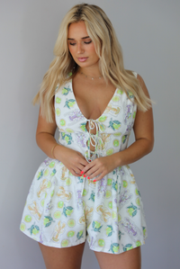 Woman wearing a white and multi-colored romper that has a lime and lobster pattern throughout, a plunging v-neckline, cut out and tie details at the bodice and waist, and slightly flowy shorts indoors against a white wall.