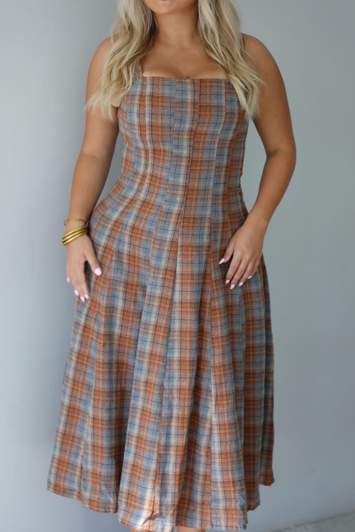 A woman wearing a brown and blue plaid midi dress with thick straps, a square neckline, fitted bodice that starts flaring out at the hips and ends about mid-shin, with a flowy skirt, standing indoors against a white wall. The photo shows a closeup of the dress, not showing the models face. 