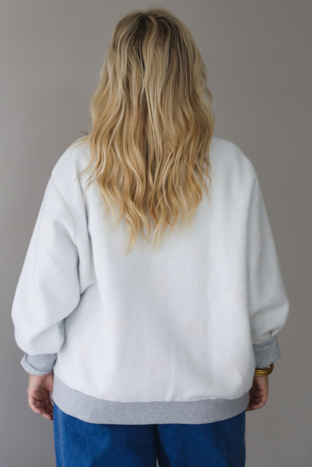 A woman shown from behind wearing a grey oversized sweatshirt with dark wash denim, indoors against a white wall. 