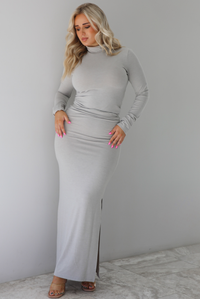 Woman wearing a grey two piece set featuring a turtle neck long sleeved fitted top and high waisted maxi length fitted skirt. The set is paired with clear strapped heels. The photo is taken indoors against a white wall with white marble flooring. 