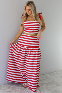 Woman wearing a red and white striped two piece set featuring a cropped top and maxi length, high waisted skirt. The photo is taken indoors against a white wall with white marble flooring. 