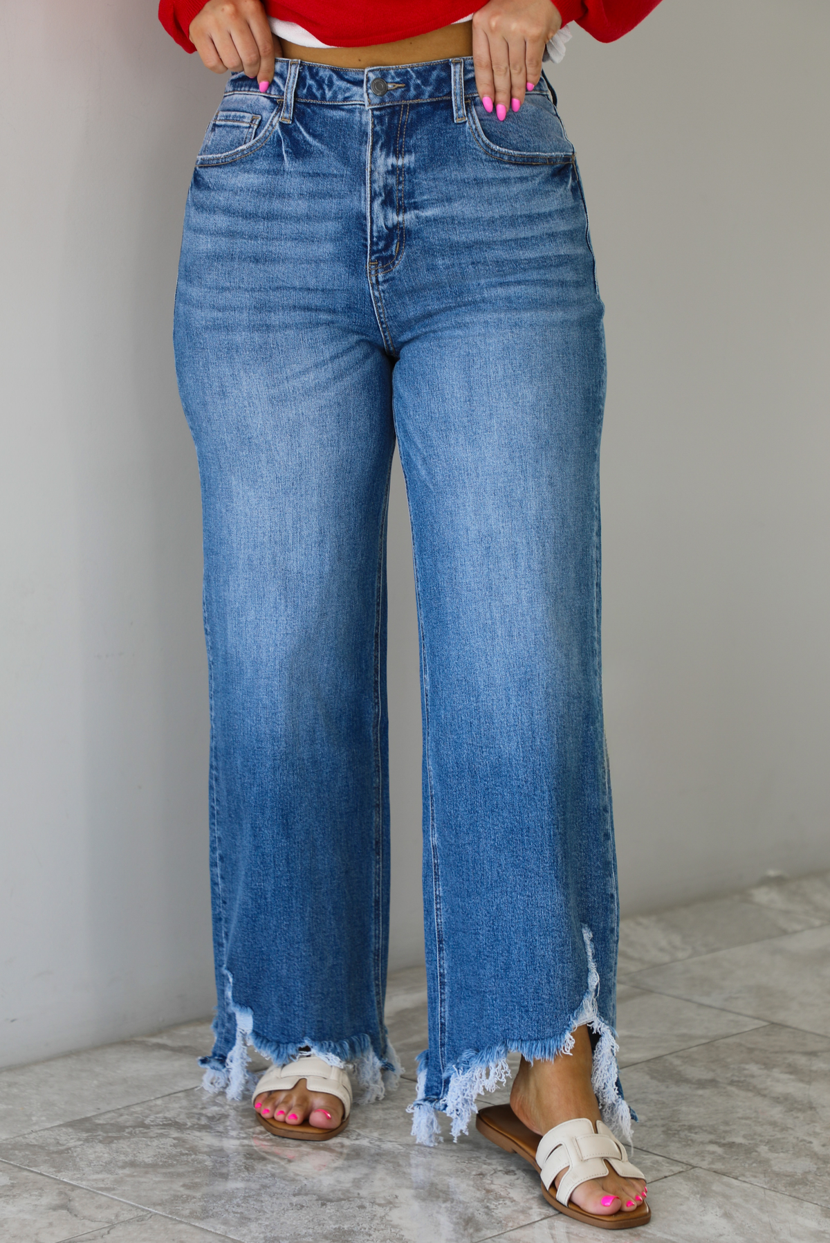Woman wearing a pair of medium wash denim that are high waisted, have functional button and zipper closures, functional front and back pockets, a straight leg design, and a raw, distressed hem. The photo is taken indoors against a white wall with white marble flooring. 