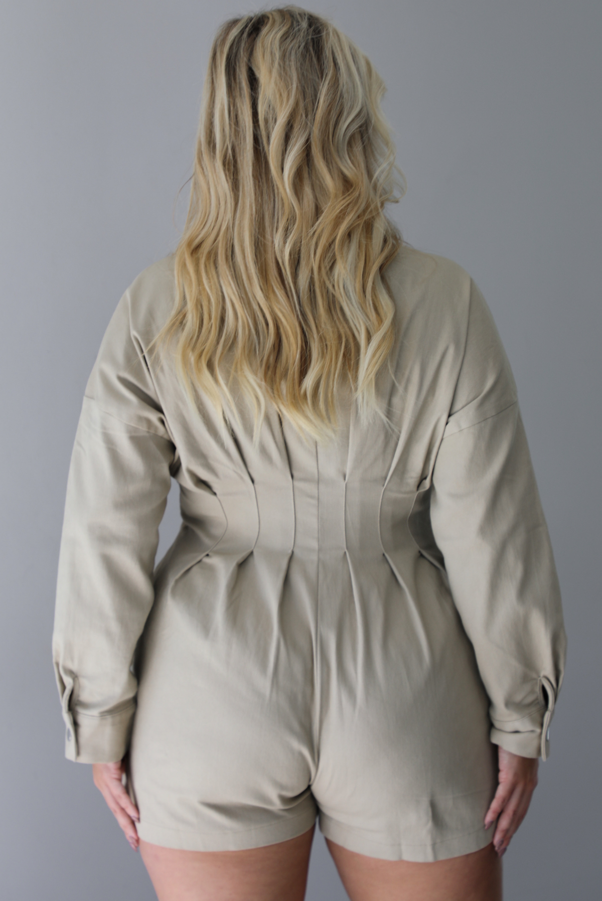 A woman shown from behind wearing a sand colored long sleeved romper with a collared neckline, functional zipper closure in the front, pleated details throughout, functional pockets on either side, standing indoors against a white wall.