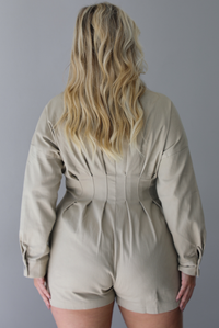 A woman shown from behind wearing a sand colored long sleeved romper with a collared neckline, functional zipper closure in the front, pleated details throughout, functional pockets on either side, standing indoors against a white wall.