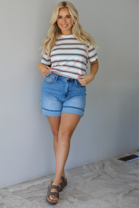 Woman wearing a striped, slightly cropped tee with light-wash denim shorts indoors against a white wall with white marble flooring. 